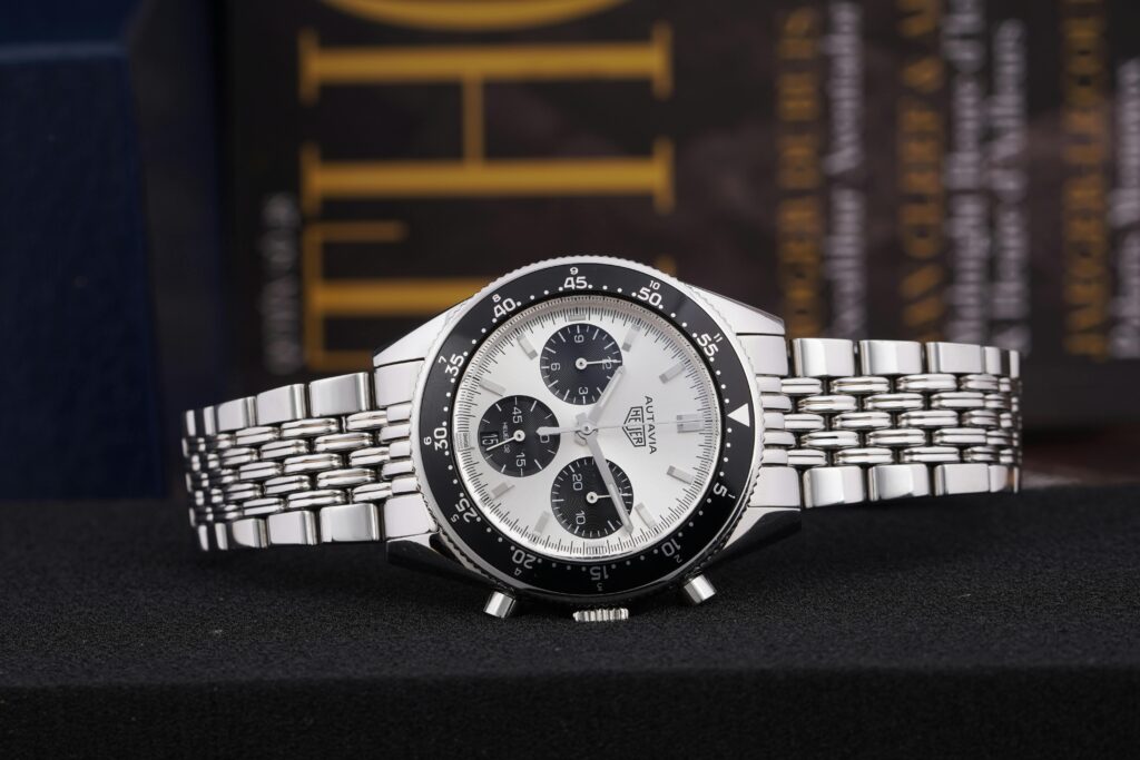 A TAG Heuer Autavia watch with a white dial, stainless steel case and bracelet, and a rotating bezel marked with tachymeter units.