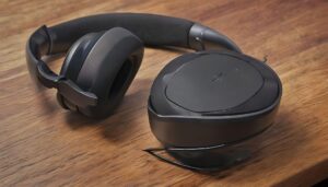 A close-up view of Sony WH-1000XM4 closed-back headphones with sleek black ear cups and headband, resting on a minimalist wooden desk with a clean, modern setup in the background. The headphones are positioned in a way that shows the soft, cushioned ear pads, with subtle lighting highlighting the matte finish."